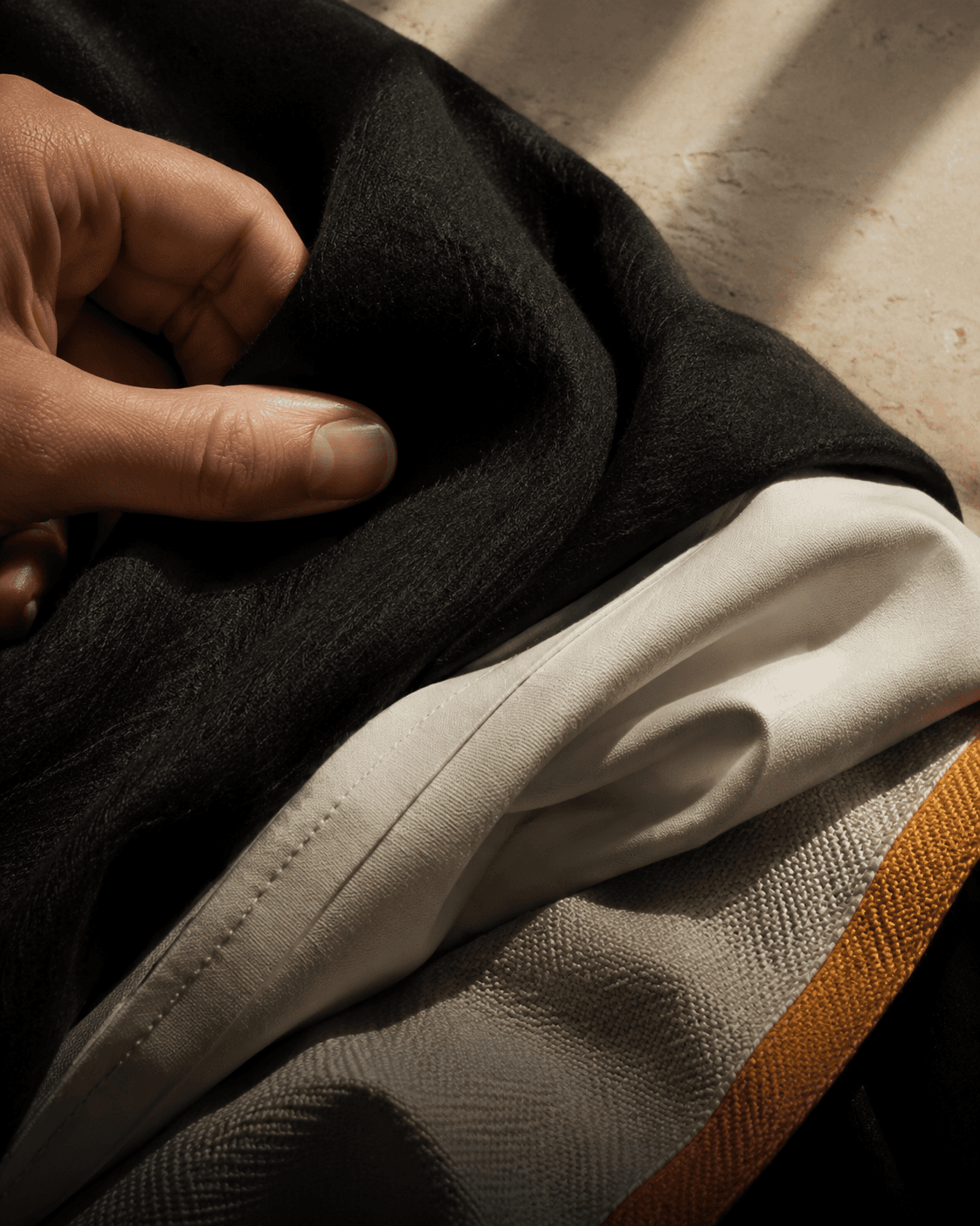 A hand feeling layered ink, bone and saffron textiles in raking daylight